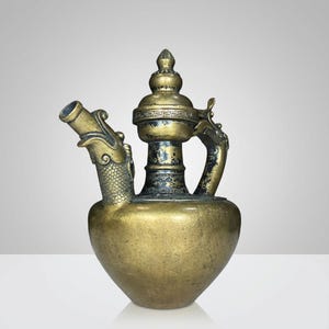 Puede incluir: Una jarra de latón vintage con una boquilla y un asa en forma de dragón. La jarra tiene un cuerpo bulboso y una tapa decorativa. El latón tiene un tono dorado con acentos más oscuros, lo que le da una apariencia antigua. La boquilla y el asa presentan intrincados detalles de dragón.