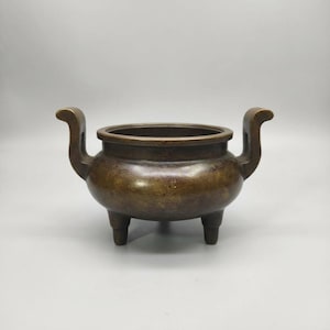 May include: A bronze-colored incense burner with a rounded body, three short legs, and two curved handles. The vessel has a wide opening and a slightly textured surface, suggesting an aged appearance. The overall design is simple and functional.