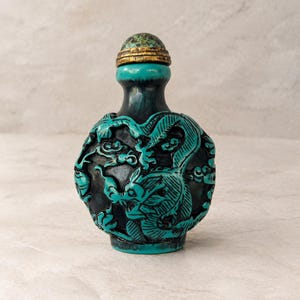 May include: A small, round snuff bottle featuring a dark base with a raised, carved turquoise-colored dragon motif wrapping around the body. The bottle is topped with a matching turquoise-colored dome-shaped stopper and a gold-toned metal rim.
