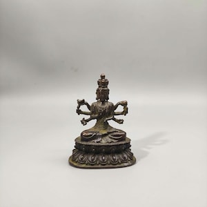Op de afbeelding: Een bronzen beeld van een godheid met meerdere armen, zittend in een meditatieve houding. Het beeld is versierd met ingewikkelde details, waaronder een kroon en een sierlijke basis. De figuur staat tegen een effen achtergrond.