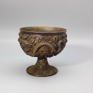 May include: Bronze-colored goblet with intricate dragon relief carvings. The cup has a wide, open bowl shape and sits on a pedestal base. The detailed design features dragons and other mythical creatures, showcasing a traditional Asian aesthetic.