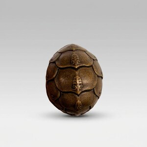 Op de afbeelding: Een bronzen, eivormig decoratief object. Het oppervlak is bedekt met overlappende, schubachtige segmenten, elk met een gedetailleerd, sierlijk ontwerp. De algehele uitstraling suggereert een ambachtelijke, antieke esthetiek.