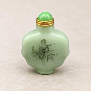 Könnte beinhalten: Eine hellgrüne Schnupftabakflasche aus Glas mit einem abgerundeten, gewellten Körper. Sie zeigt eine schwarze Strichzeichnung einer sitzenden Figur, die eine Schriftrolle hält. Die Flasche hat einen goldfarbenen Metallkragen und einen grünen Stopfen.