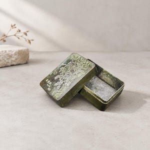 May include: A rectangular metal box with a weathered green finish and a hinged lid. The lid features an embossed landscape illustration of trees and structures, accompanied by small characters. The interior of the box is metallic and silver.