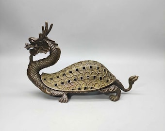 May include: A bronze-colored dragon turtle figurine. The dragon has a detailed head with antlers and a curved neck. The turtle shell is covered in coin-shaped cutouts. The figurine is on a white background.