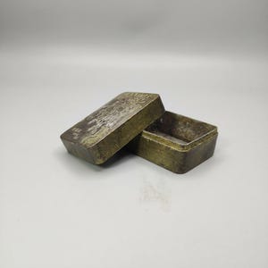 May include: A rectangular metal box with a hinged lid, slightly ajar. The box has an aged, tarnished appearance with etched designs on the lid. The box is likely used for storing small items.