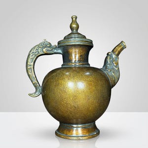 Puede incluir: Tetera antigua de bronce con cuerpo redondo, tapa y boquilla. El asa y la boquilla están adornadas con motivos de dragones. La tetera es de color bronce oscuro con una ligera pátina, lo que sugiere antigüedad. Se apoya sobre una pequeña base redonda.