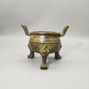 Peut inclure: Un brûleur d'encens à trois pieds de couleur bronze avec deux poignées. Le récipient a un corps rond avec des sculptures décoratives et un design détaillé sur les pieds. L'esthétique générale suggère une pièce antique ou vintage.