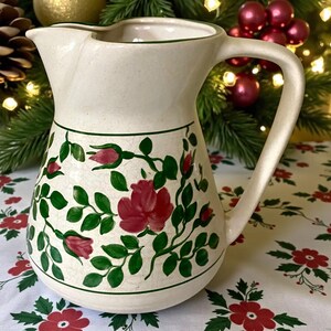 Peut inclure: Pichet en céramique crème avec une anse et un bec courbés. Le pichet est décoré de roses rouges et de feuilles vertes, soulignées d'une bordure verte. Le motif floral enveloppe le corps du pichet.