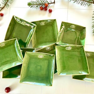 Può includere: Una collezione di piatti quadrati in ceramica verde con un emblema decorativo al centro. I piatti hanno una finitura lucida e una consistenza leggermente irregolare. I piatti sono disposti su una superficie bianca, con rametti di verde e bacche rosse.