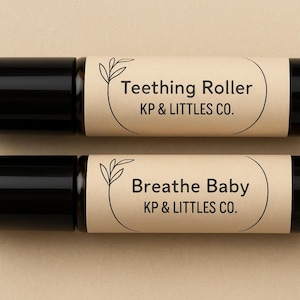 Puede incluir: Dos botellas de rodillo negras con etiquetas color canela. La botella superior dice "Teething Roller" y la inferior "Breathe Baby". Ambas etiquetas incluyen el texto "KP & LITTLES CO." y un diseño de hojas.