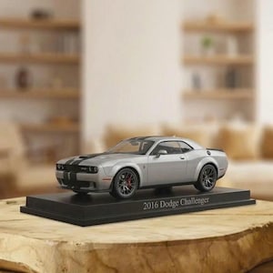 May include: A silver 2016 Dodge Challenger scale model car featuring black racing stripes on the hood and roof. The vehicle is mounted on a black rectangular display base with the text 2016 Dodge Challenger printed in white on the front edge.