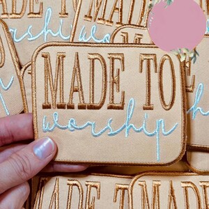 May include: Tan embroidered patch with the words "MADE TO worship" in gold and light blue thread. The patch has a rectangular shape with rounded corners and a gold border. Several patches are visible.