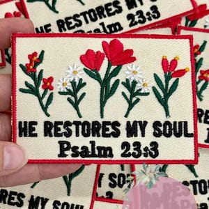 May include: Embroidered patch with the text "HE RESTORES MY SOUL Psalm 23:3" in black. The patch features embroidered red and white flowers with green stems on a cream background, framed by a red border.