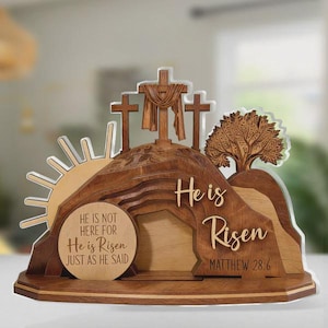 Puede incluir: Decoración de Pascua de madera con un amanecer, tres cruces y un árbol. Se muestra el texto "He is Risen" y un versículo bíblico. El diseño incluye una tumba con las palabras "He is not here for He is Risen just as He said."