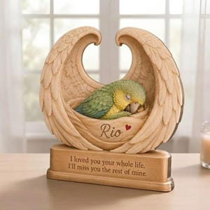 May include: A tan-colored resin sculpture featuring a green and yellow parrot nestled within a pair of carved angel wings. The base displays the name Rio with a small red heart and the engraved text: I loved you your whole life, I'll miss you the rest of mine.