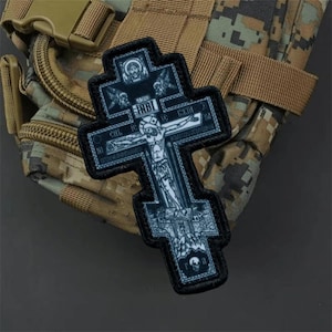 May include: A black and white embroidered patch in the shape of an Eastern Orthodox cross. The design features a detailed depiction of the crucifixion of Jesus with the letters INBI at the top, surrounded by religious iconography and a skull.