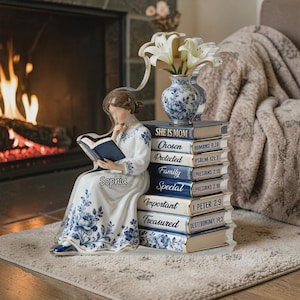 Puede incluir: Figura de porcelana de una mujer leyendo un libro, sentada junto a una pila de libros y un jarrón con flores. La figura lleva un vestido blanco con motivos florales azules. Los libros tienen títulos como "She is Mom" y "Treasured".