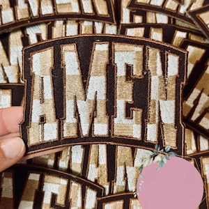 May include: Embroidered patch with the word "AMEN" in a bold, block letter style. The letters are white and tan, with a checkered pattern, set against a dark brown background. The patch has a brown border.