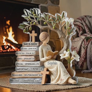 Puede incluir: Figura decorativa de una mujer leyendo, junto a una pila de libros con inscripciones religiosas. También hay un olivo y un jarrón con flores. La figura incluye el nombre "Sophia".