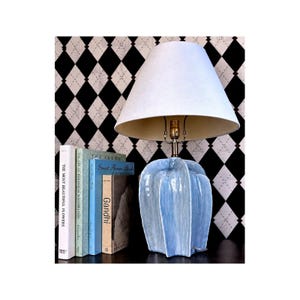 May include: A table lamp with a light blue, vertically ribbed ceramic base and a plain white conical fabric lampshade. The lamp is positioned next to a row of four books with visible spines, including titles like The Most Beautiful Flowers and Gandhi.