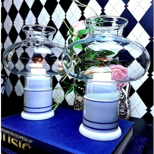 May include: Two glass candle holders with a bulbous top and a cylindrical base. The base is white with blue stripes. A lit candle sits inside each holder, and pink flowers are visible in the background.