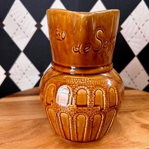 May include: A brown ceramic pitcher with a wide, flared top and a rounded base. The pitcher features embossed detailing, including an arched design around the base and text on the upper portion. The pitcher is displayed on a wooden surface.