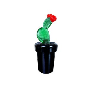 May include: A glass sculpture of a green cactus with two bright red flowers on top, set into a solid black cylindrical base. The cactus features a textured, translucent surface, while the base has a smooth, glossy, and opaque finish.