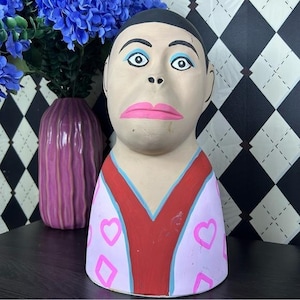May include: A hand-painted ceramic bust with a stylized face, featuring pink lips, blue eye shadow, and a red and pink patterned top. The bust is displayed near a purple vase and blue flowers, against a black and white diamond pattern background.