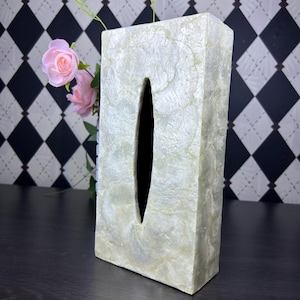 May include: Rectangular tissue box cover with a pearlescent finish. The cover has a vertical oval opening and is set against a black and white diamond pattern background. Pink roses are visible in the background.