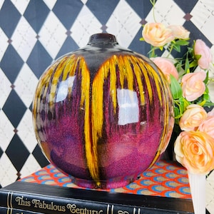 May include: A spherical ceramic vase with a glossy finish. The vase features a gradient of colors, transitioning from a dark brown top to vibrant yellow and purple hues. The vase is sitting on a stack of books.