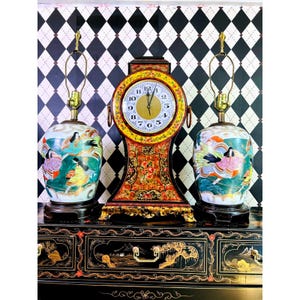 May include: A decorative black cabinet with gold accents supports a vintage clock and two matching lamps. The clock has a gold face and a red and gold frame. The lamps have a white base with a colorful Asian-inspired design.
