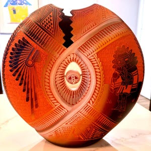 May include: A round, orange and black ceramic vase with a central circular cutout containing a small sphere. The surface features intricate etched geometric patterns, a profile of a person wearing a feathered headdress, and a figure in ceremonial attire.