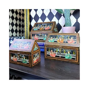 May include: Three rectangular wooden boxes with hinged, gabled lids. Each box features hand-painted scenes of figures in traditional attire on a dark background, framed by intricate gold and black borders. The boxes have metal latches on the front.
