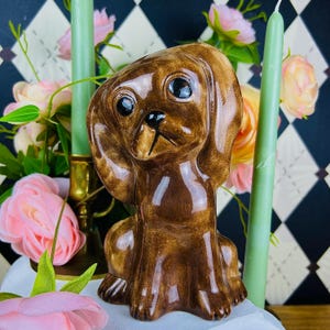 May include: A brown ceramic dog figurine with large, expressive eyes. The dog is sitting upright with its ears flopped down. The figurine is surrounded by pink roses, green candles, and a gold candle holder.