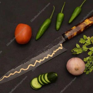 Puede incluir: Un cuchillo de chef con un patr&oacute;n de onda dorada decorativa en la hoja, junto con ingredientes frescos: un tomate, pimientos verdes, un pepino en rodajas, una cebolla y cilantro. El cuchillo tiene un mango marr&oacute;n estampado.