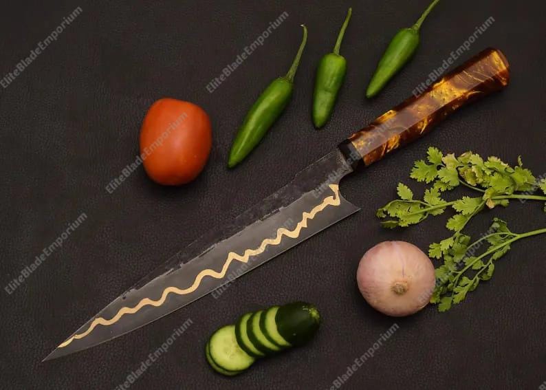 Puede incluir: Un cuchillo de chef con un patr&oacute;n de onda dorada decorativa en la hoja, junto con ingredientes frescos: un tomate, pimientos verdes, un pepino en rodajas, una cebolla y cilantro. El cuchillo tiene un mango marr&oacute;n estampado.