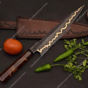 Puede incluir: Cuchillo de chef artesanal con una hoja oscura y un dise&ntilde;o ondulado dorado. Se muestra con una funda de cuero, un tomate, una chalota, chiles verdes y cilantro. Ideal para la cocina.