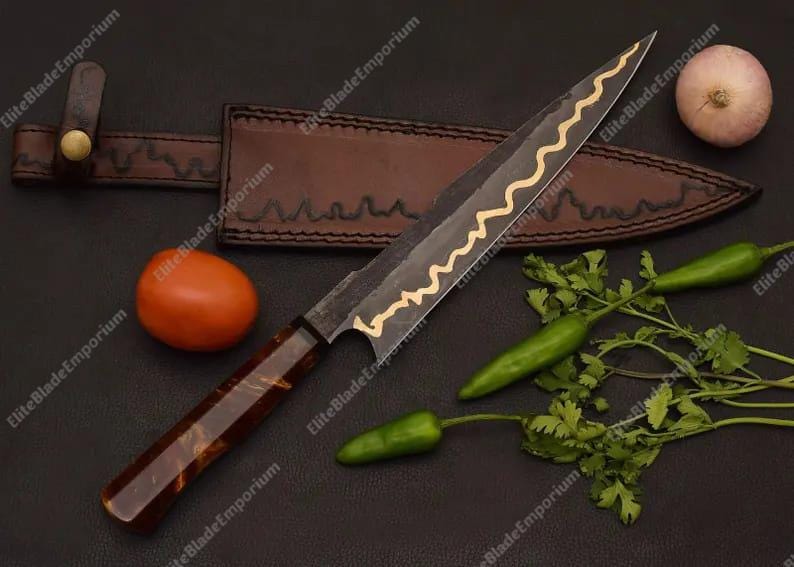 Puede incluir: Cuchillo de chef artesanal con una hoja oscura y un dise&ntilde;o ondulado dorado. Se muestra con una funda de cuero, un tomate, una chalota, chiles verdes y cilantro. Ideal para la cocina.