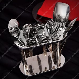 Könnte beinhalten: Ein Edelstahl-Besteckset, präsentiert in einem silberfarbenen Halter. Das Set umfasst Löffel, Gabeln und Messer, die zur Präsentation angeordnet sind. Der Halter hat ein schlankes, modernes Design mit schwarzem Rand.