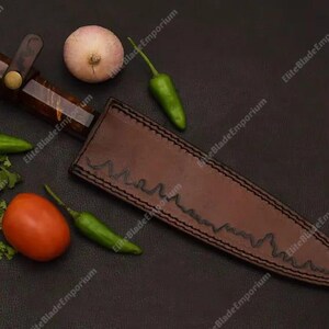 Puede incluir: Un cuchillo artesanal con mango marr&oacute;n y funda de cuero. La imagen tambi&eacute;n presenta verduras frescas, como un tomate, pimientos verdes y una cebolla. El cuchillo y la funda est&aacute;n sobre una superficie oscura.