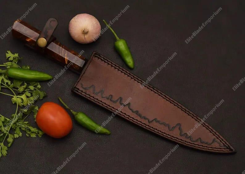 Puede incluir: Un cuchillo artesanal con mango marr&oacute;n y funda de cuero. La imagen tambi&eacute;n presenta verduras frescas, como un tomate, pimientos verdes y una cebolla. El cuchillo y la funda est&aacute;n sobre una superficie oscura.