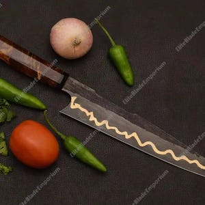 Puede incluir: Un cuchillo de chef artesanal con un mango de madera oscura y una hoja con un dise&ntilde;o decorativo dorado. El cuchillo est&aacute; rodeado de ingredientes frescos: un tomate, una cebolla, chiles verdes y cilantro, todo sobre una superficie oscura.