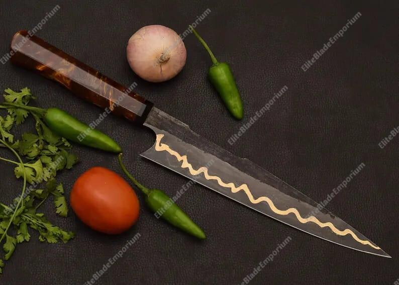 Puede incluir: Un cuchillo de chef artesanal con un mango de madera oscura y una hoja con un dise&ntilde;o decorativo dorado. El cuchillo est&aacute; rodeado de ingredientes frescos: un tomate, una cebolla, chiles verdes y cilantro, todo sobre una superficie oscura.