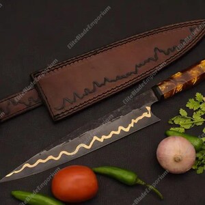 Puede incluir: Un cuchillo de chef artesanal con una funda de cuero marr&oacute;n. El cuchillo tiene una hoja oscura con un dise&ntilde;o ondulado dorado y un mango estampado. Verduras frescas, incluyendo un tomate, cebolla y pimientos, est&aacute;n dispuestas alrededor del cuchillo.