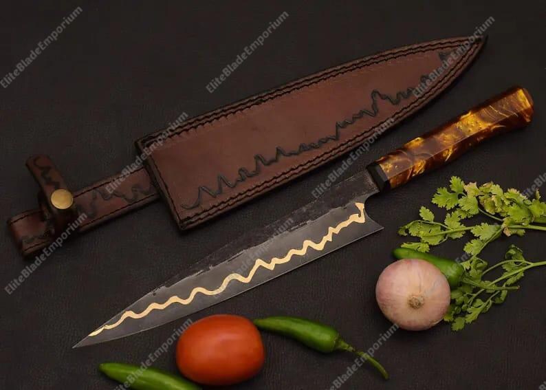 Puede incluir: Un cuchillo de chef artesanal con una funda de cuero marr&oacute;n. El cuchillo tiene una hoja oscura con un dise&ntilde;o ondulado dorado y un mango estampado. Verduras frescas, incluyendo un tomate, cebolla y pimientos, est&aacute;n dispuestas alrededor del cuchillo.
