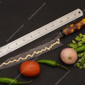 Puede incluir: Un cuchillo de chef largo y negro con un dise&ntilde;o dorado decorativo a lo largo de la hoja, junto a una regla. El cuchillo tiene un mango marr&oacute;n y dorado. Tambi&eacute;n hay verduras frescas, como un tomate, cebolla y pimientos.