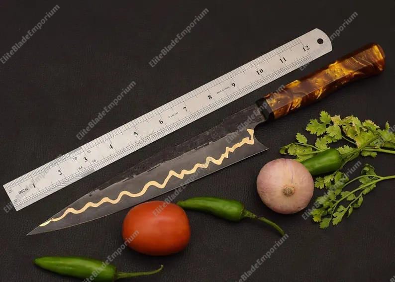 Puede incluir: Un cuchillo de chef largo y negro con un dise&ntilde;o dorado decorativo a lo largo de la hoja, junto a una regla. El cuchillo tiene un mango marr&oacute;n y dorado. Tambi&eacute;n hay verduras frescas, como un tomate, cebolla y pimientos.
