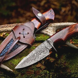 Puede incluir: Un cuchillo de caza artesanal con una hoja de acero estampada y un mango de madera, junto a su funda de cuero marr&oacute;n. La funda presenta un dise&ntilde;o de cruz decorativa y una trabilla para el cintur&oacute;n. El cuchillo y la funda est&aacute;n sobre musgo y madera.