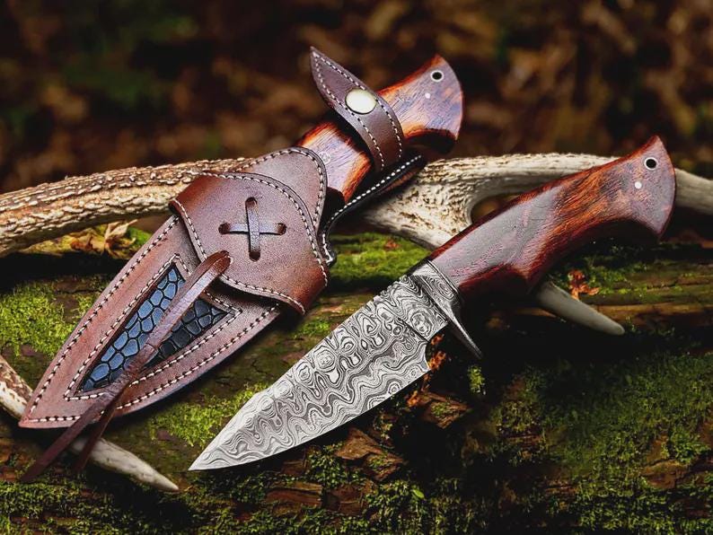 Puede incluir: Un cuchillo de caza artesanal con una hoja de acero estampada y un mango de madera, junto a su funda de cuero marr&oacute;n. La funda presenta un dise&ntilde;o de cruz decorativa y una trabilla para el cintur&oacute;n. El cuchillo y la funda est&aacute;n sobre musgo y madera.